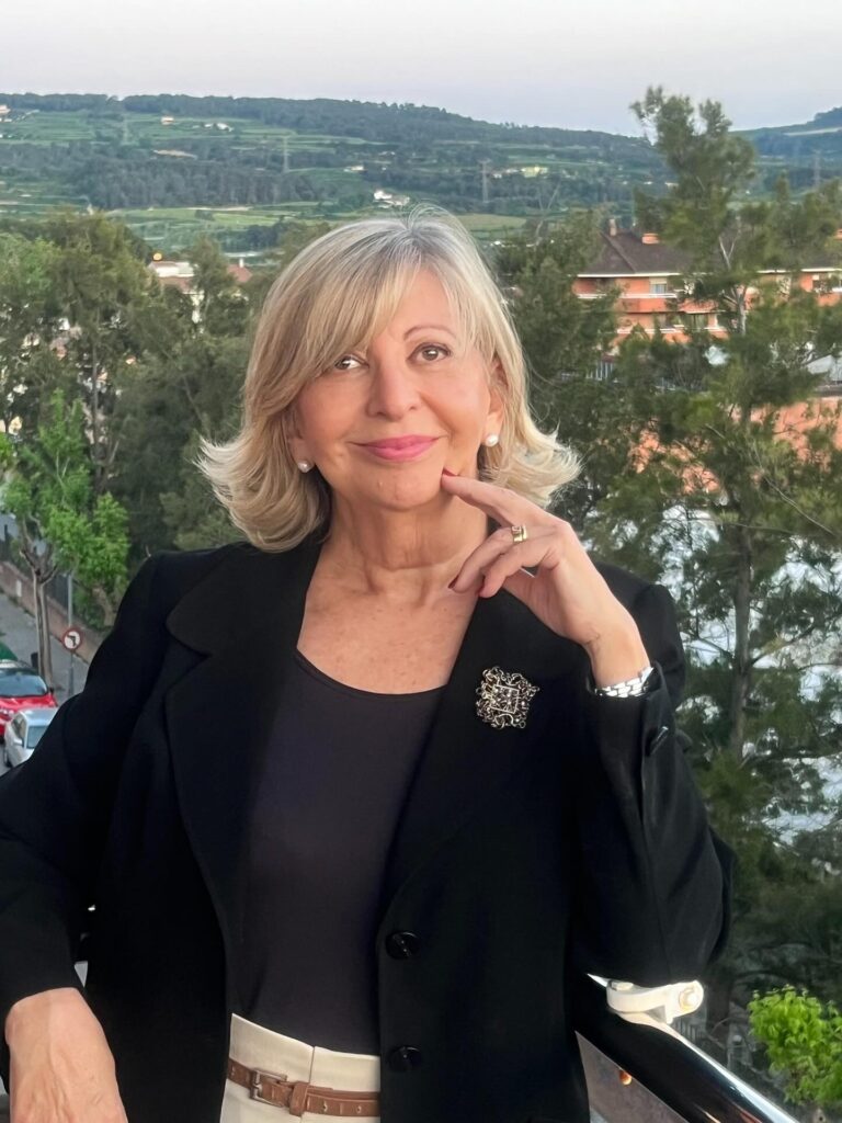 A woman over 60 with a round face wearing a mid-length blonde hairstyle with flicked-out ends and side-swept bangs.