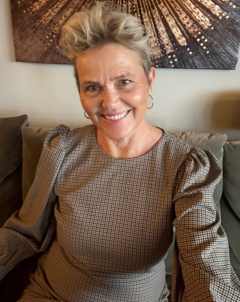 A smiling woman over 60 with a round face and a very short, textured blonde pixie cut featuring a tall, voluminous quiff on top.