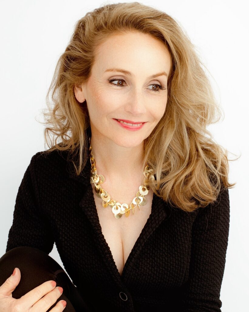 A woman over 60 with a round face wearing shoulder-length honey-blonde hair with voluminous swept-back layers and soft waves.