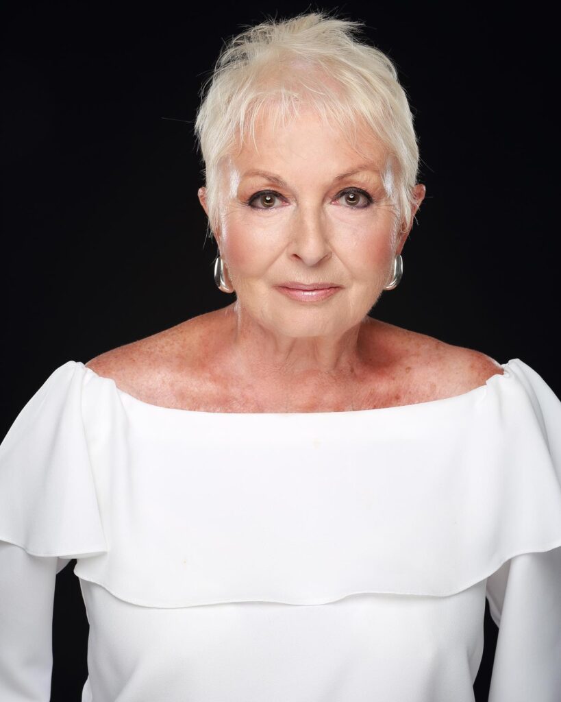 A woman over 60 with a round face wearing a short wispy platinum blonde pixie haircut with tapered sides.