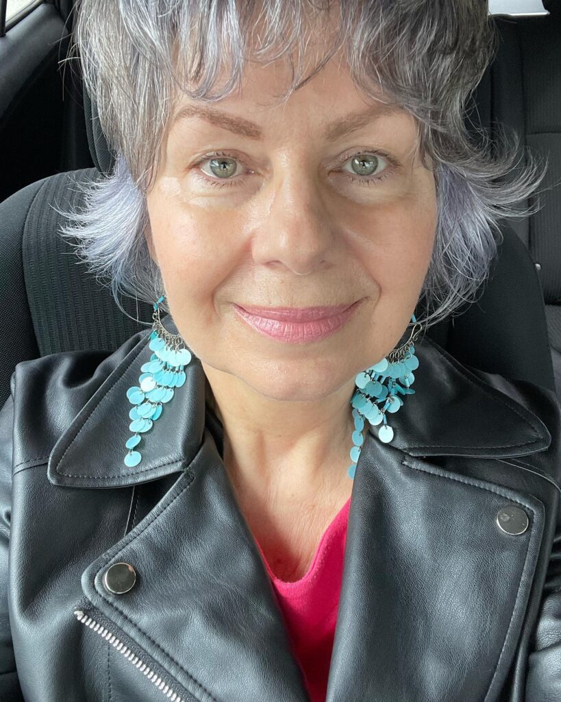 A woman over 60 with a round face wearing a textured silver and lavender pixie cut with flared side layers.