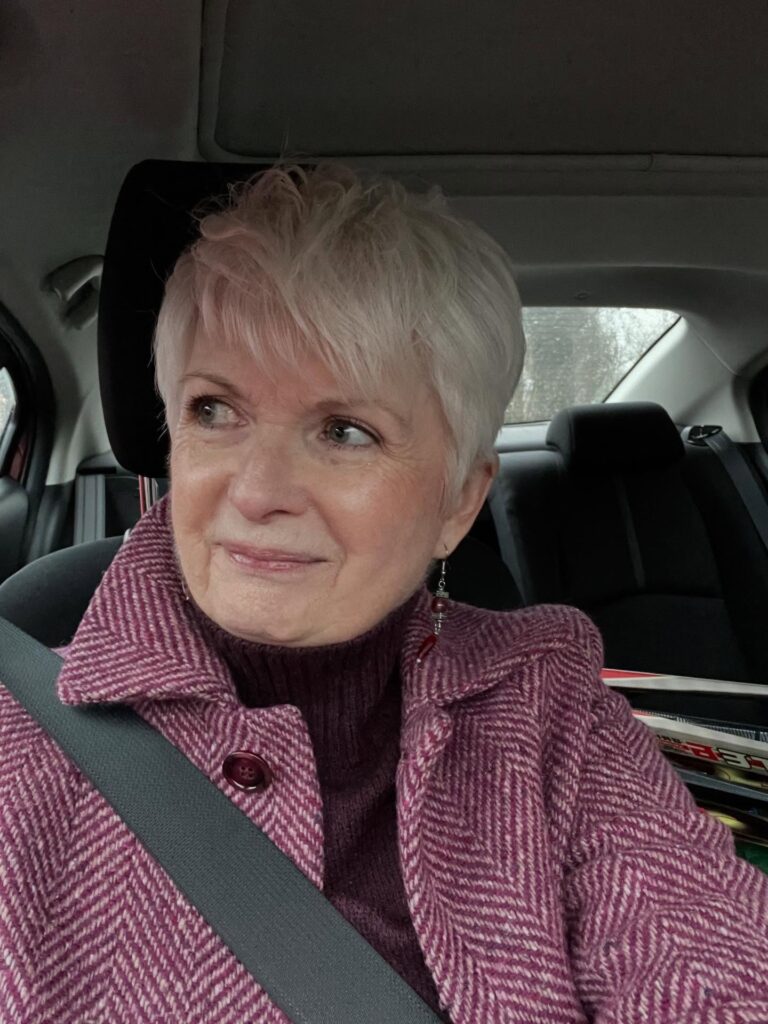 A woman over 60 with a round face wearing a short platinum pixie cut with wispy layers and soft bangs.
