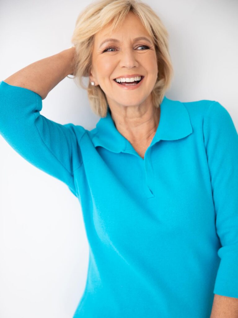 A smiling woman over 60 with a round face wearing a voluminous blonde layered bob hairstyle.