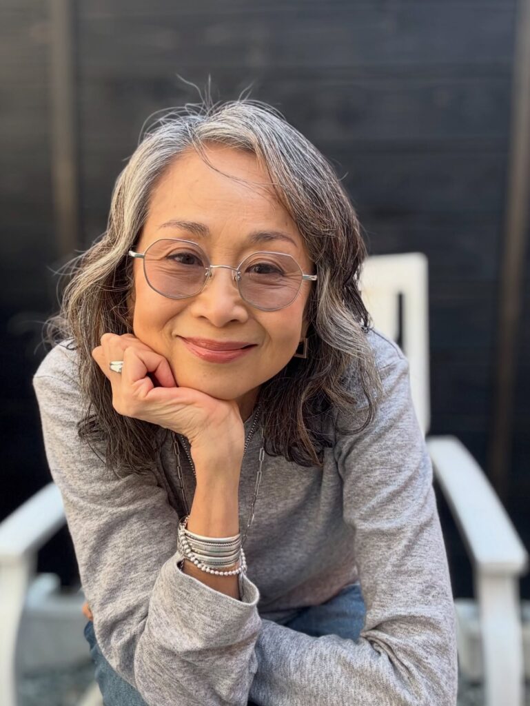 A woman over 60 with a round face wearing a wavy shoulder-length salt-and-pepper hairstyle with a side part.