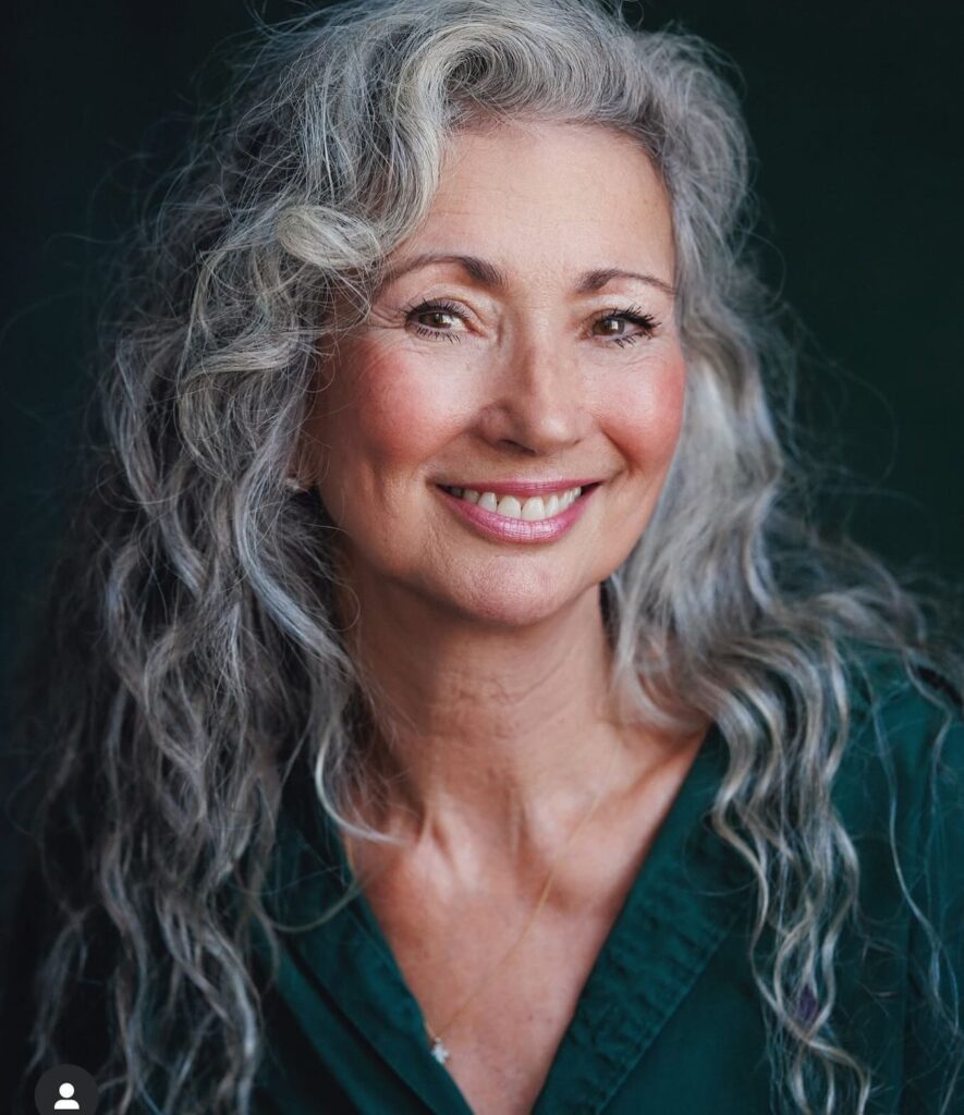A smiling woman over 60 with a round face wearing long, voluminous silver curls with layered texture.