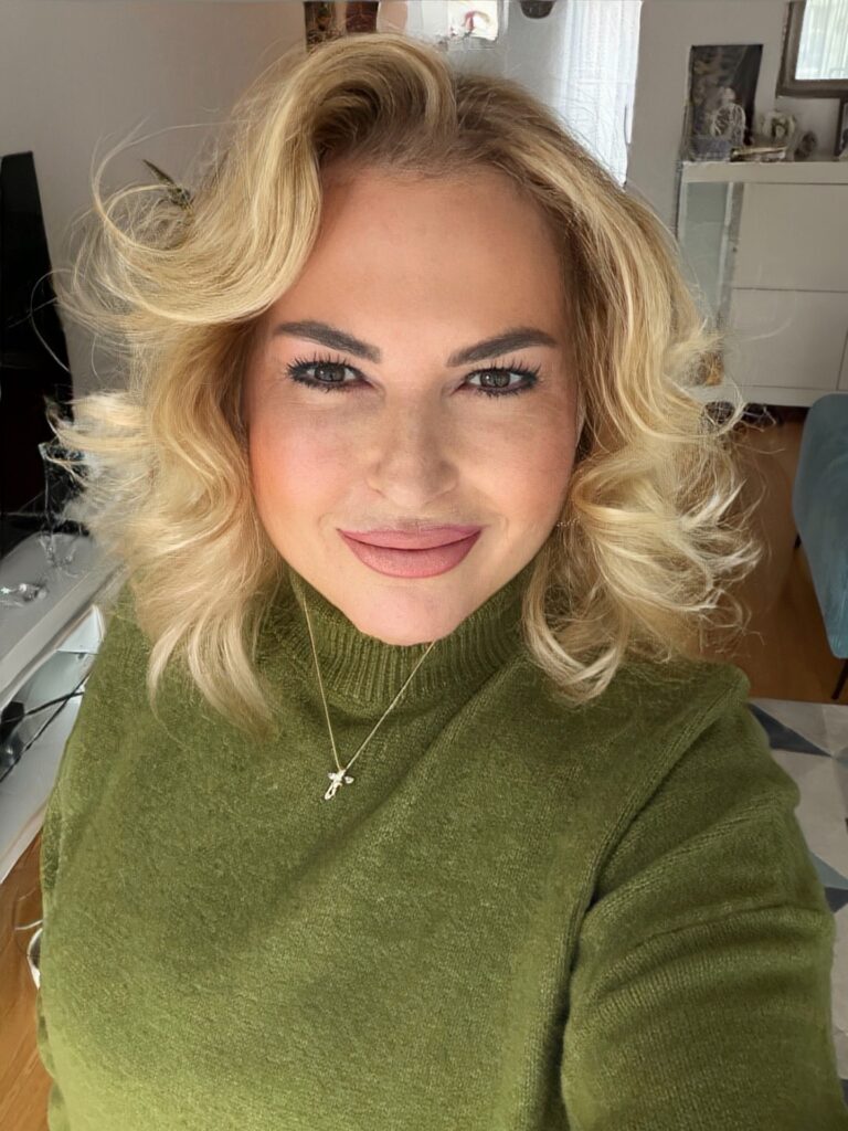 A woman over 60 with a round face wearing voluminous golden-blonde shoulder-length curls with a deep side part.