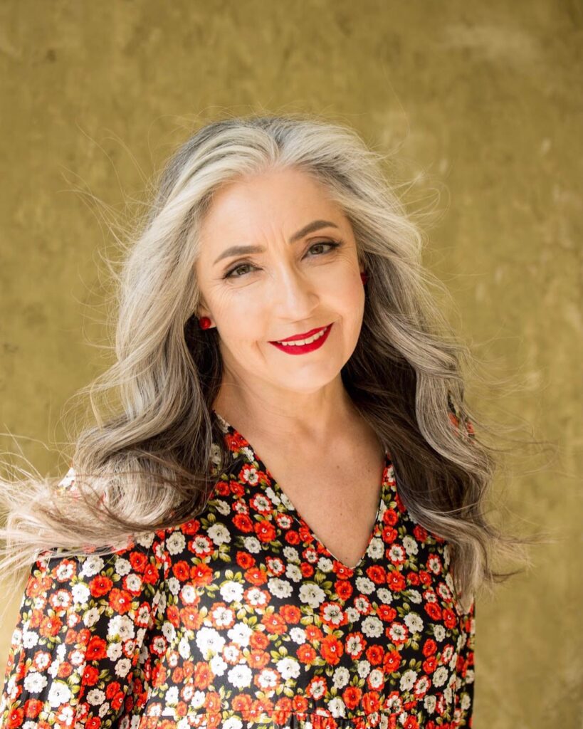 A smiling woman over 60 with a round face wearing long wavy silver hair with bright white highlights around her face.