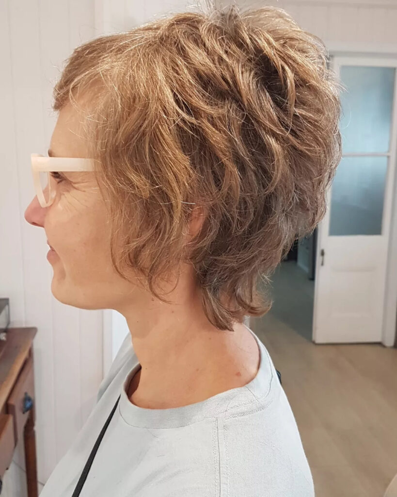 A woman in profile wearing a tousled honey-brown pixie cut with soft waves and voluminous layers.