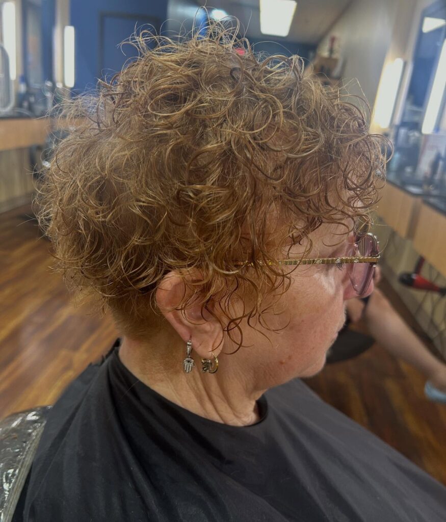 A woman over 60 with a short curly pixie haircut in a warm honey blonde color featuring tight natural ringlets.
