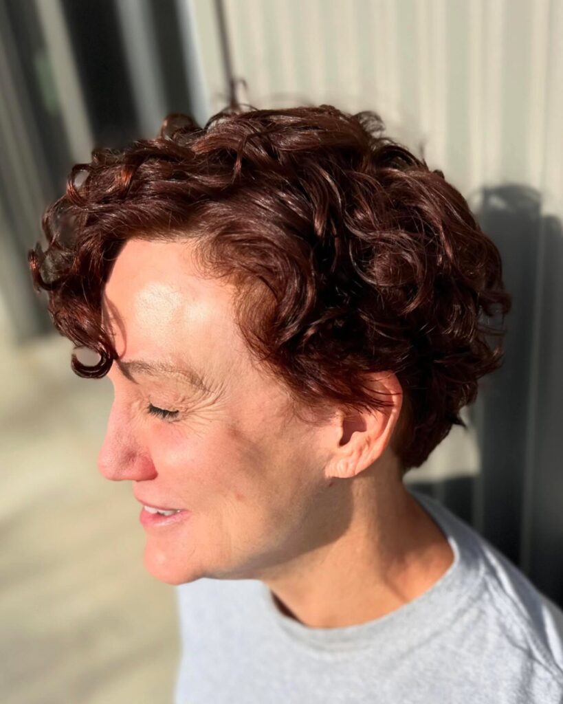 A side profile of a woman over 60 with a glossy mahogany red curly pixie cut featuring loose, defined waves.