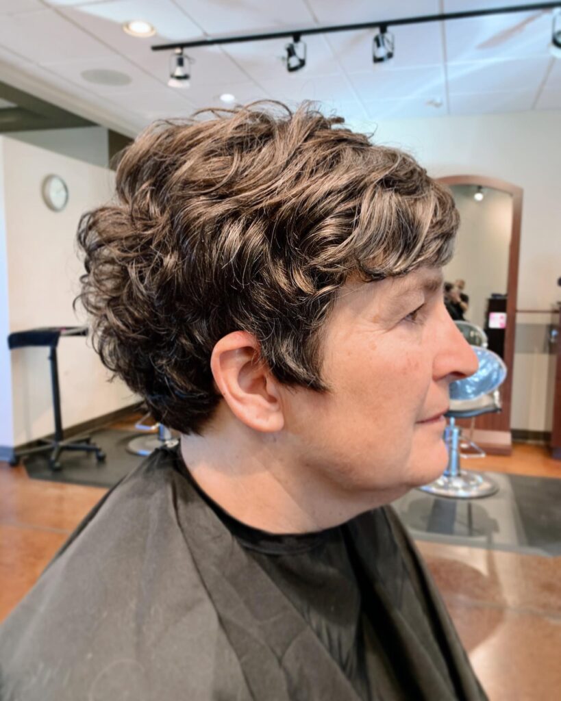 A side profile of a woman over 60 with a layered wavy pixie cut in cool ash brown tones.