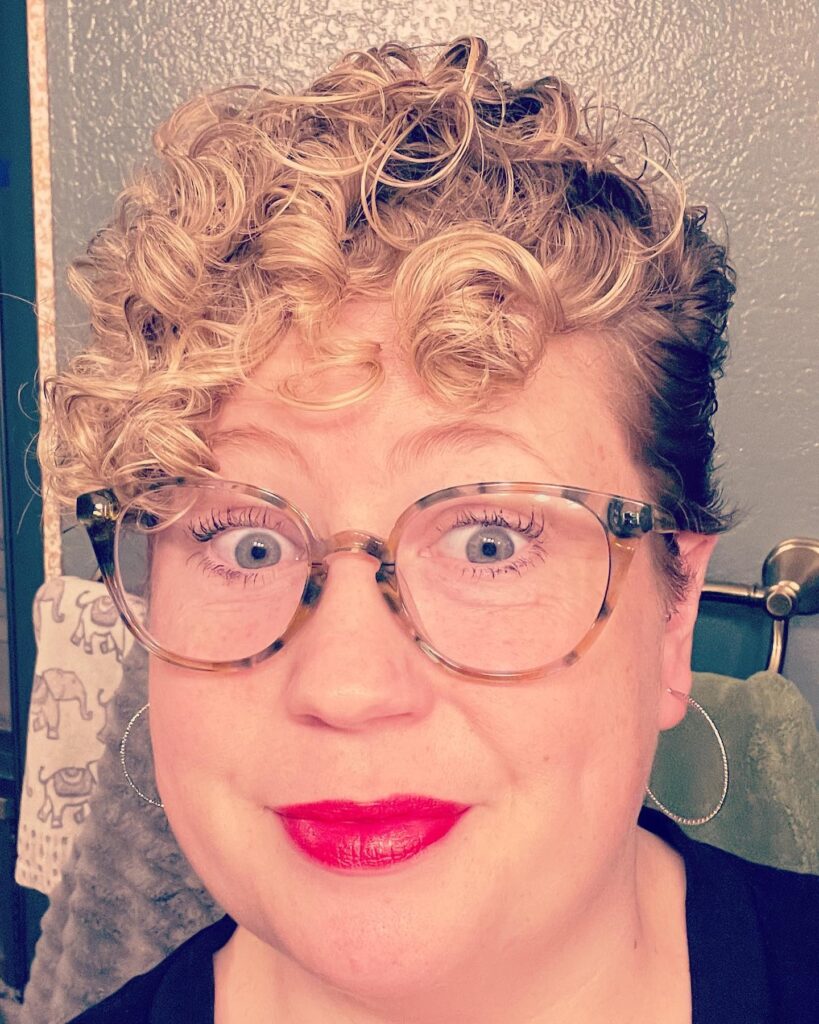 A woman over 60 with a golden blonde pixie haircut featuring voluminous tight curls styled upward and forward on top.