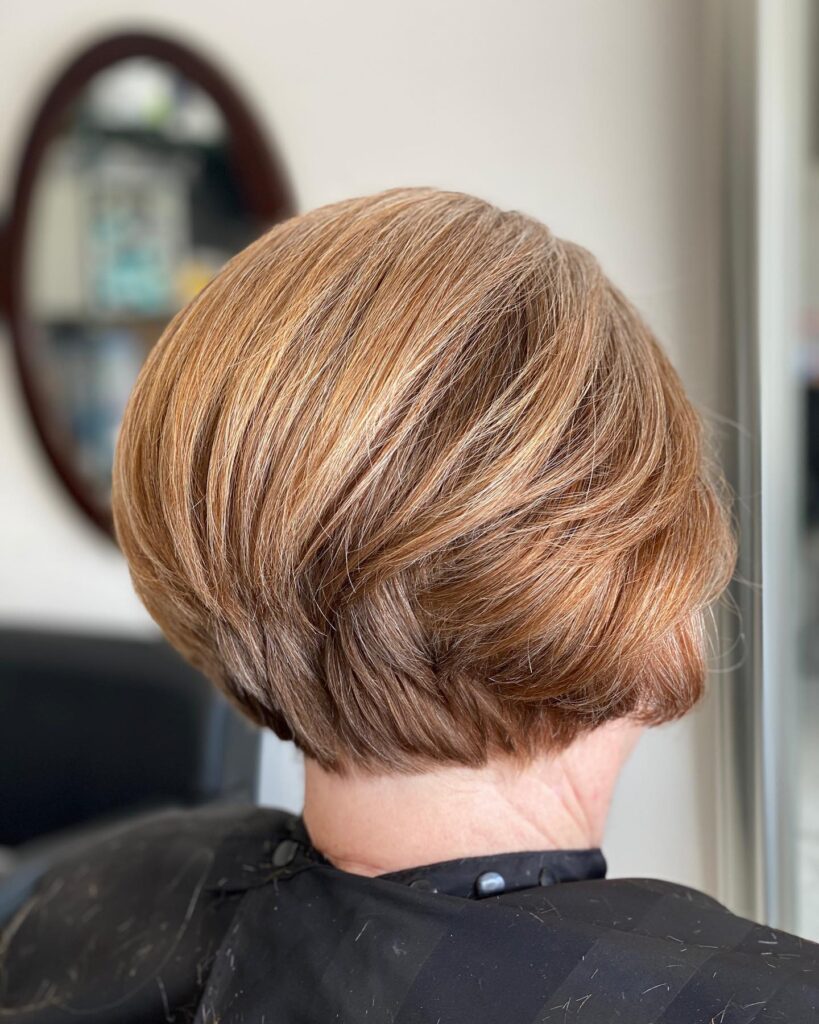 A woman with a warm, honey-blonde stacked bob haircut showing textured layers and volume at the back of the head.