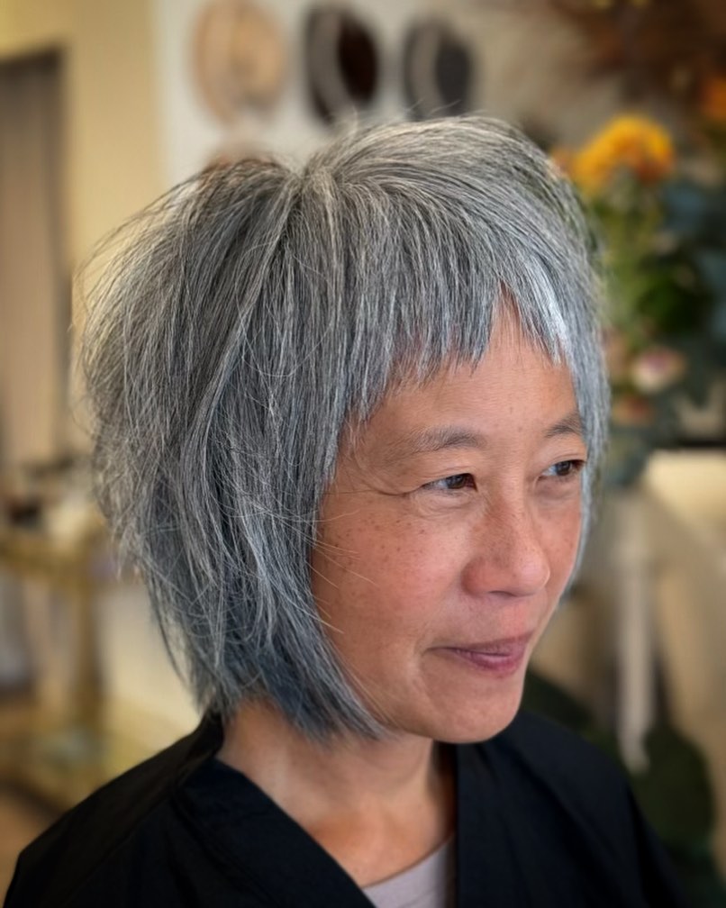 A woman with a short choppy steel-grey bob featuring a jagged micro fringe and a messy textured finish.