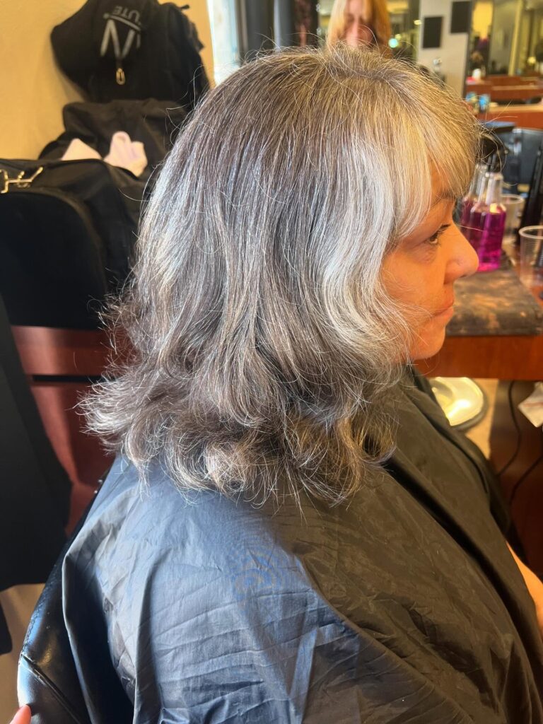 A woman with a medium-length salt-and-pepper layered haircut featuring wavy ends and full wispy bangs.