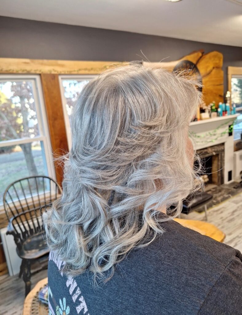 A woman viewed from the back showing a medium-length silver hairstyle with voluminous curls and tapered layers.