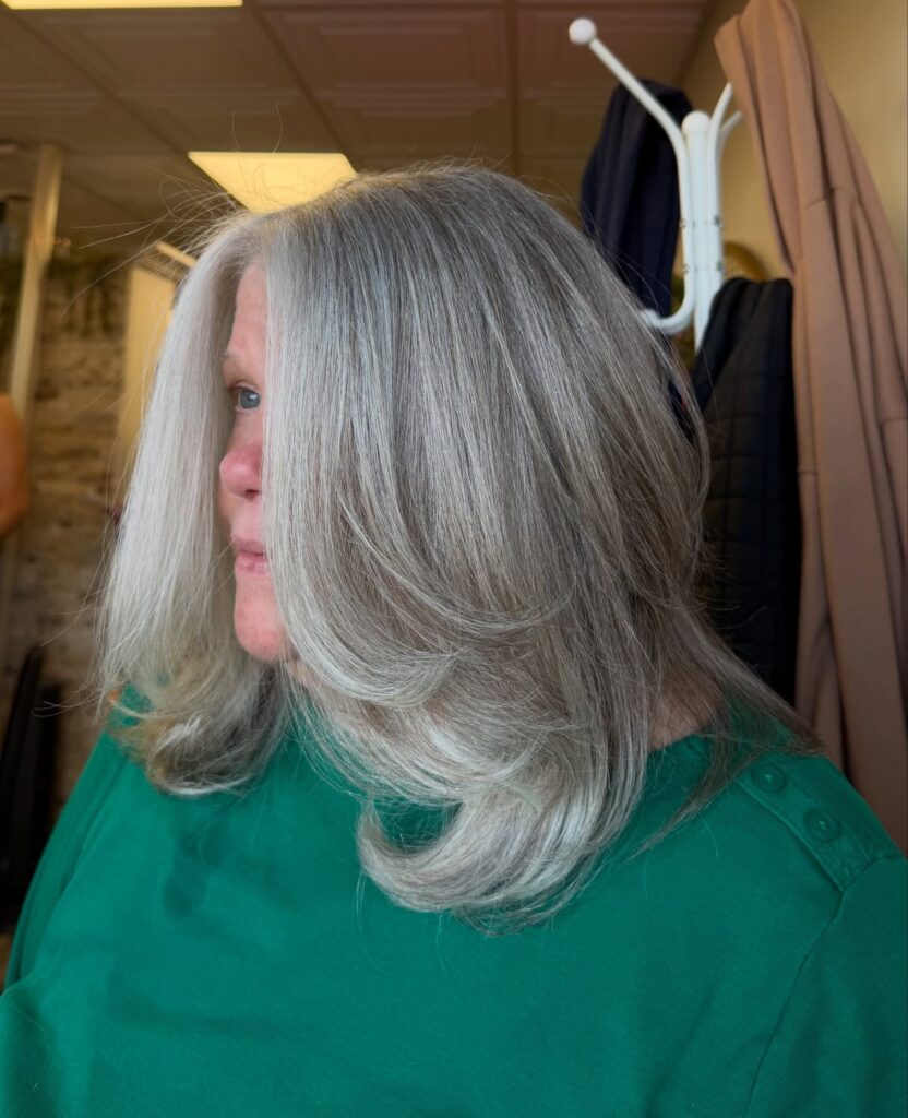 A woman with a medium-length silver layered haircut featuring long side-sweeping layers and ends flipped outward.