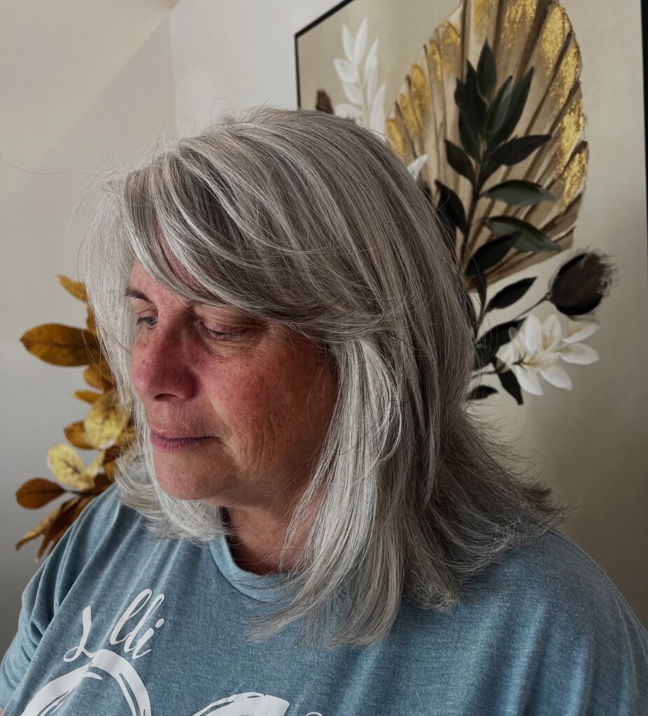 A woman with a medium-length salt-and-pepper layered haircut featuring side-swept bangs and smooth, straight layers.
