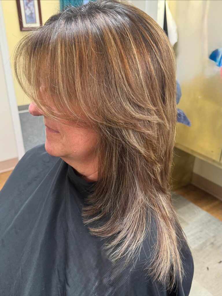 A woman with a medium-length honey brown layered haircut featuring golden highlights and soft side-swept bangs.