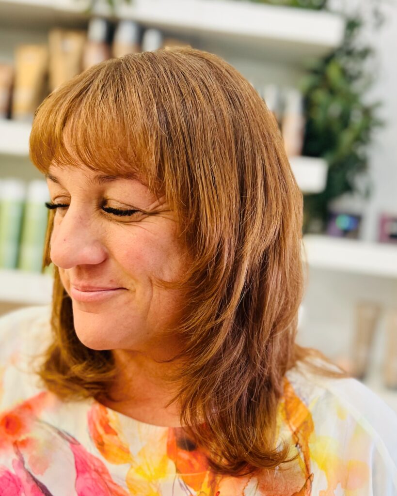 A woman with a medium-length auburn layered haircut featuring an inward-curving butterfly cut and full bangs.