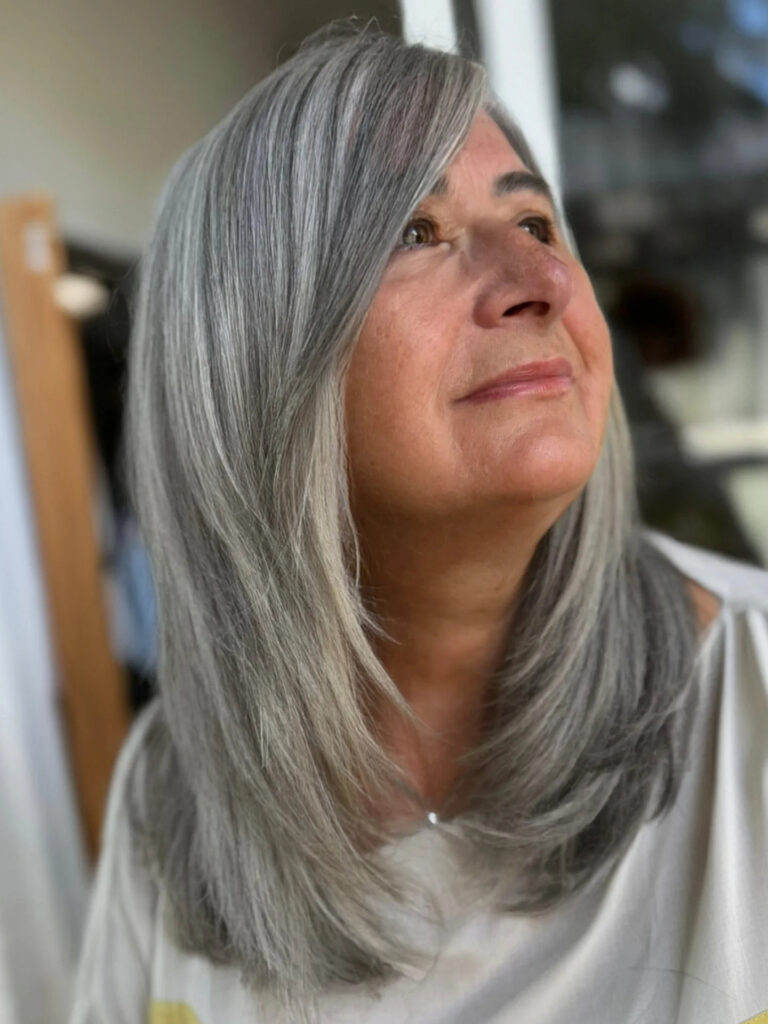 A woman with a medium-length silver layered haircut featuring a side part and long cascading layers that curve toward the face.