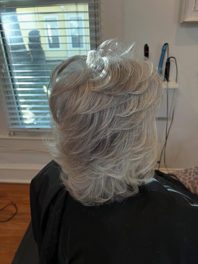 A back view of a woman with a medium-length ash blonde haircut showing multiple stacked layers and voluminous styling.