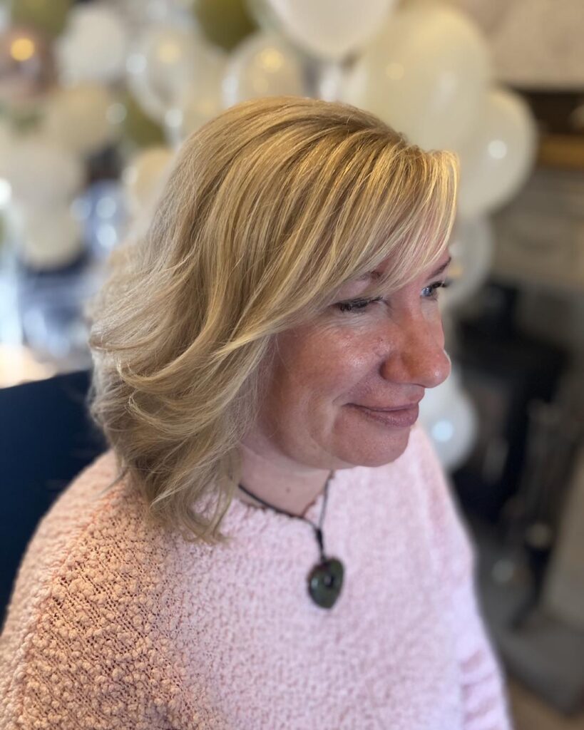 A woman with a medium-length honey-blonde layered bob featuring side-swept bangs and soft textured layers.