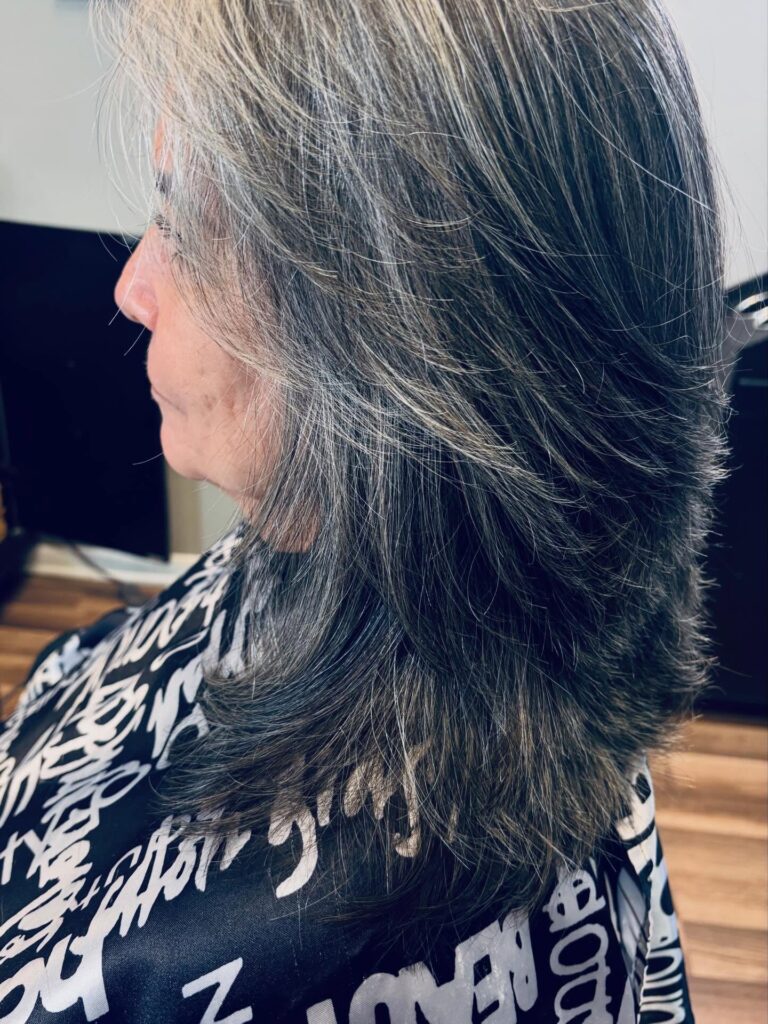 A woman with a medium length salt and pepper layered haircut featuring distinct stepped layers and feathered ends.