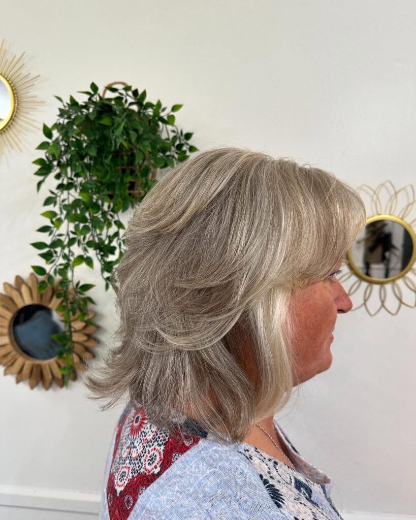A woman from the side with a medium-length ash-blonde layered haircut featuring flicked-out ends and a soft fringe.