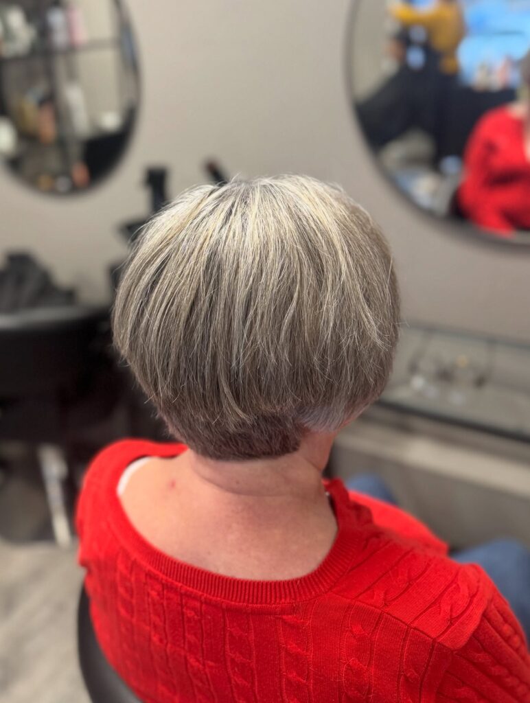 A back view of a woman with a rounded blonde-and-grey bob featuring a short buzzed undercut at the nape of the neck.