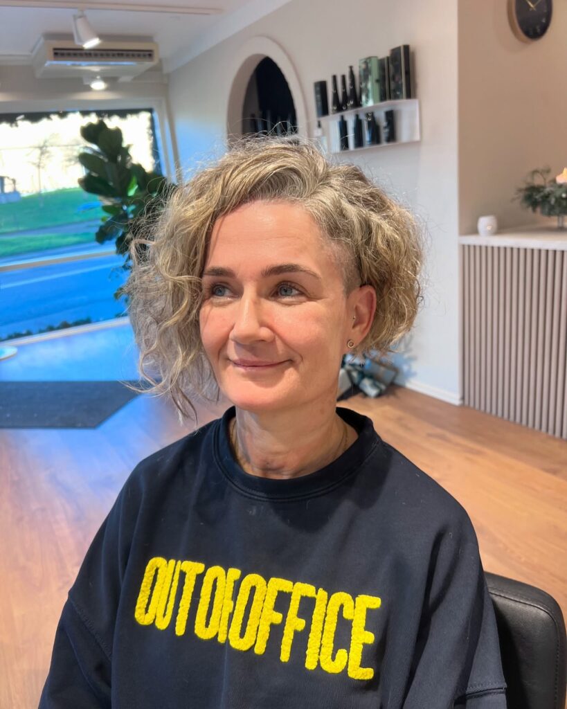 A woman over 60 with an asymmetrical curly bob featuring a side undercut and sandy ash-blonde tones.