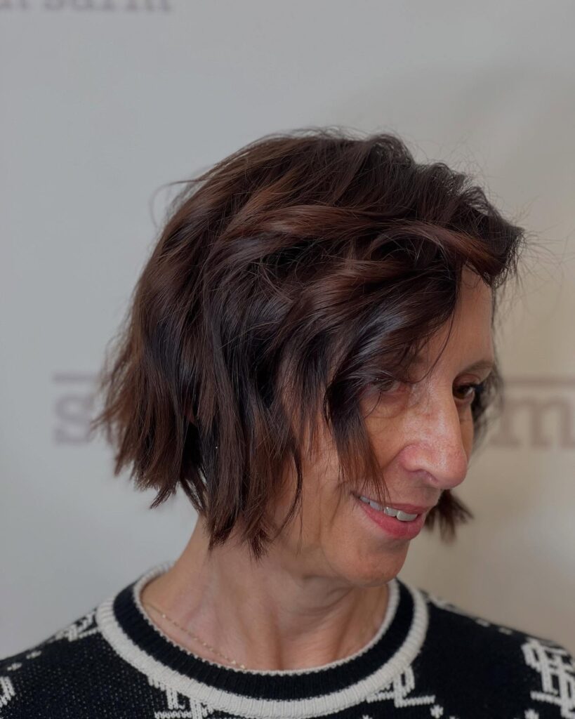 A woman over 60 with a textured mocha-brunette bob featuring choppy ends and soft, messy waves.
