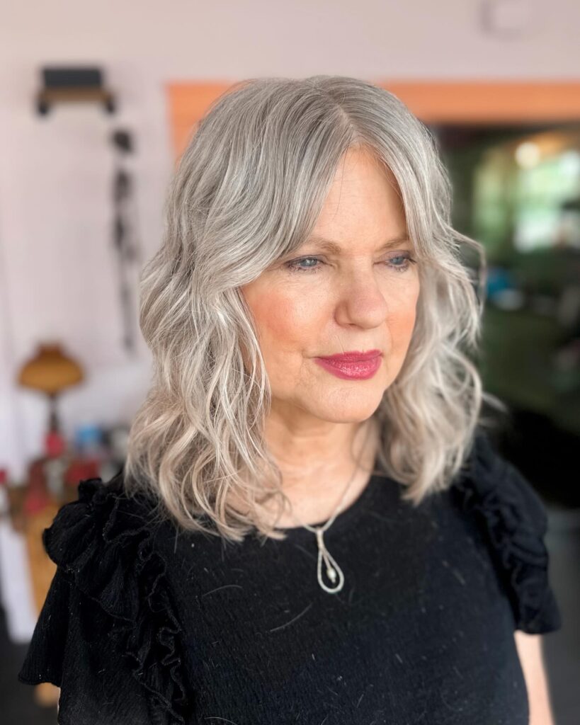 An older woman with shoulder-length wavy cool silver hair and curtain bangs, wearing a black top.