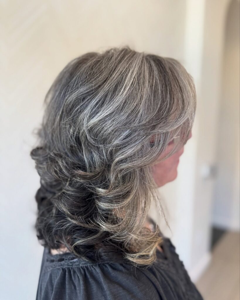 A side profile of a woman with medium-length voluminous feathered hair in a mix of dark grey and silver tones.