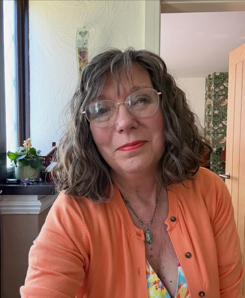 A woman with shoulder-length, wavy, ash-grey hair and soft bangs, wearing glasses and an orange cardigan.