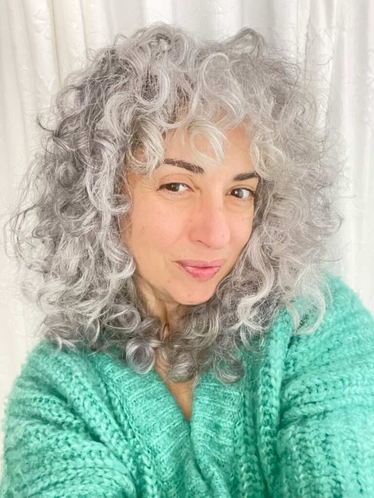 A woman with shoulder-length voluminous curly grey hair and curly bangs wearing a teal sweater.