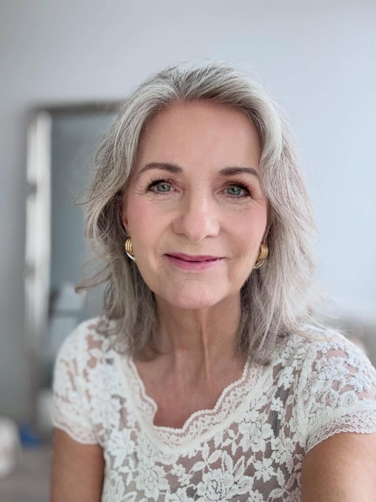 A woman over sixty with shoulder-length wavy grey hair and a gentle side part, wearing a white lace top.