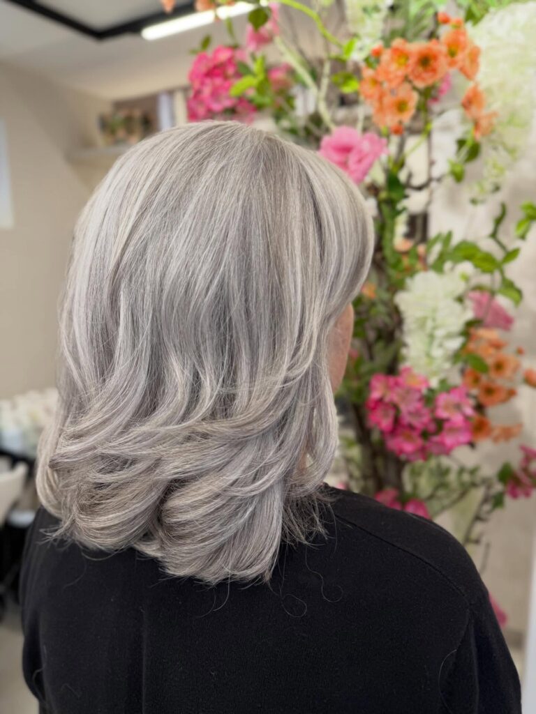 A back view of a woman with medium-length bright silver hair featuring long layers and curled-out ends.