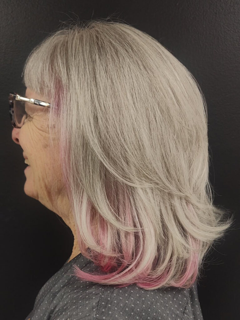 A side profile of a woman with a medium-length grey haircut featuring straight bangs and bright pink highlights underneath.
