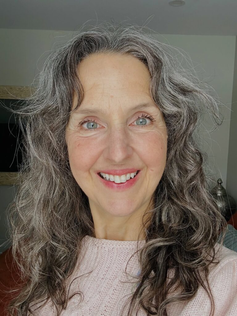 A smiling woman with medium-length curly salt-and-pepper hair and a soft side part wearing a pink sweater.