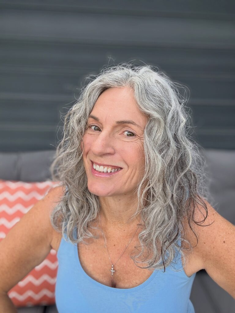 A smiling woman with medium-length wavy grey hair and a center part, wearing a blue tank top.
