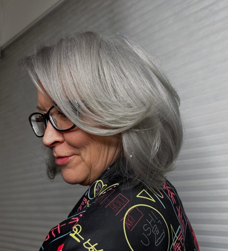 A woman wearing glasses with a medium-length silver bob that features long face-framing layers and ends that curve inward toward the jawline.