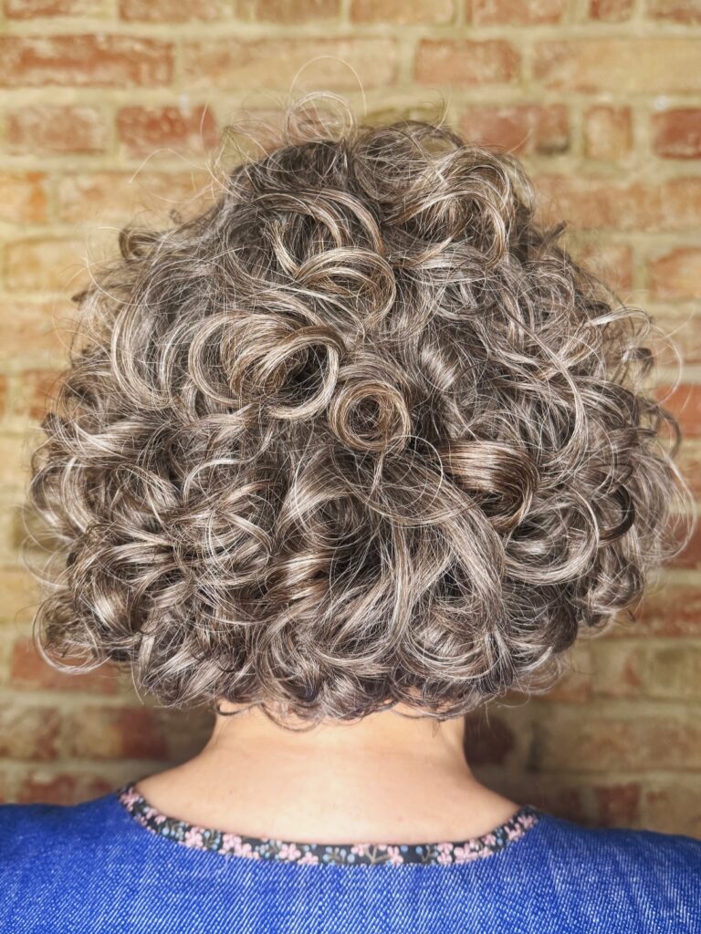 A woman viewed from behind with a voluminous medium-length curly bob in a salt and pepper brunette shade.