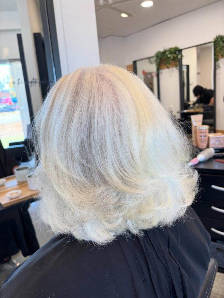A woman viewed from behind with a voluminous medium-length layered bob in a bright platinum blonde shade featuring flipped ends.