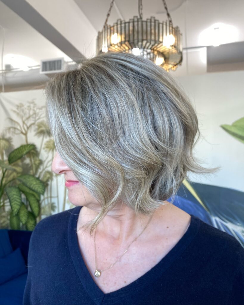 A woman in profile with a tousled medium bob featuring ash blonde and gray tones with beachy waves and piecey layers.