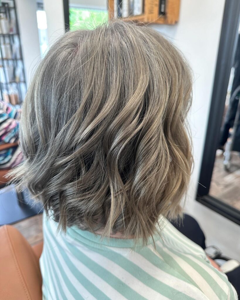 A woman viewed from behind with a medium-length wavy bob in mushroom blonde with ashy highlights and choppy ends.