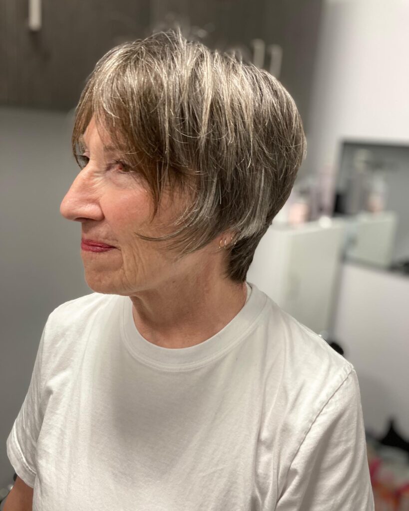 A side profile of a woman over 60 with a textured gray pixie bob featuring wispy bangs and soft feathered layers.