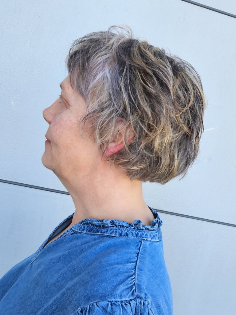 A side view of a woman with a wavy salt-and-pepper bixie cut featuring honey blonde highlights and a voluminous back.