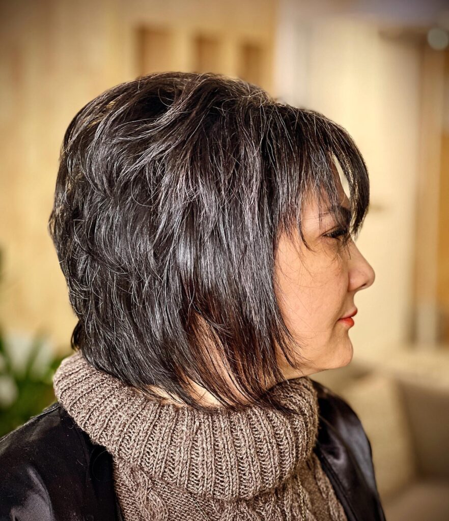 A side profile of a woman over 60 with a voluminous dark charcoal pixie bob featuring shaggy layers and silver highlights.