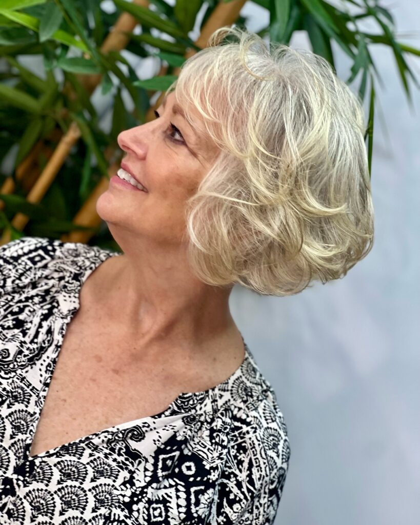 A side profile of a smiling woman with a voluminous platinum blonde bixie cut featuring soft romantic waves and airy bangs.